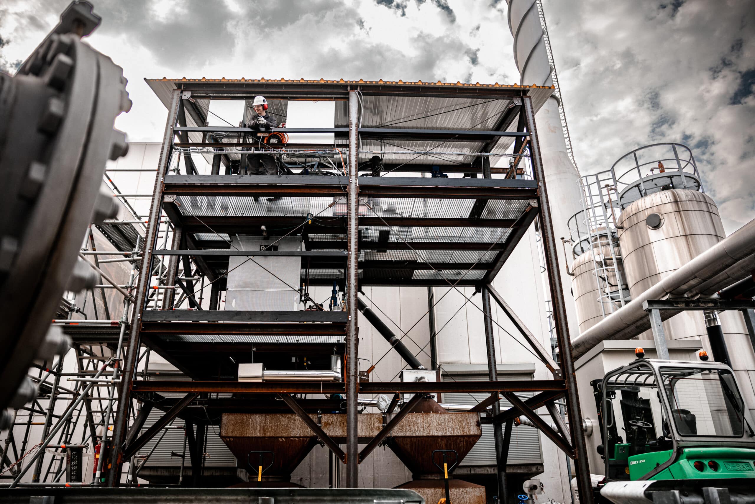 A worker wearing safety gear stands on the second level of a metal industrial structure at a factory, with machinery, pipes, and storage tanks visible under a cloudy sky.