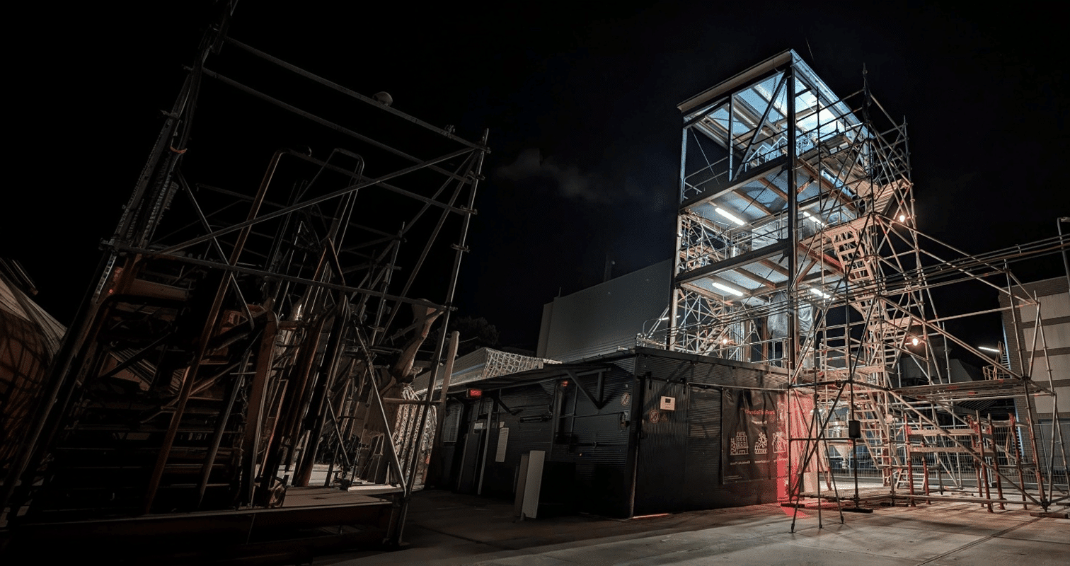 A tall, multi-level scaffolding structure is illuminated at night, surrounded by more metal scaffolding and construction equipment against a dark sky.