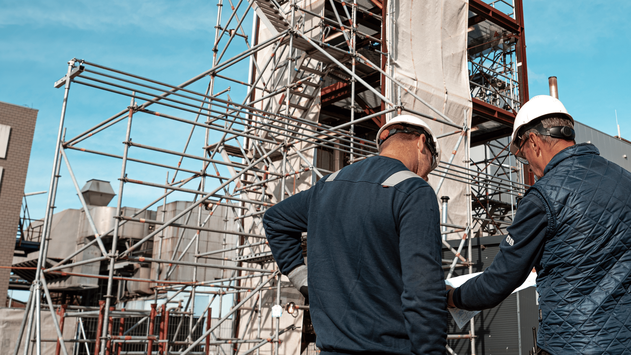 Two construction workers wearing hard hats review plans at a building site, surrounded by scaffolding and partially completed structures under a clear blue sky.