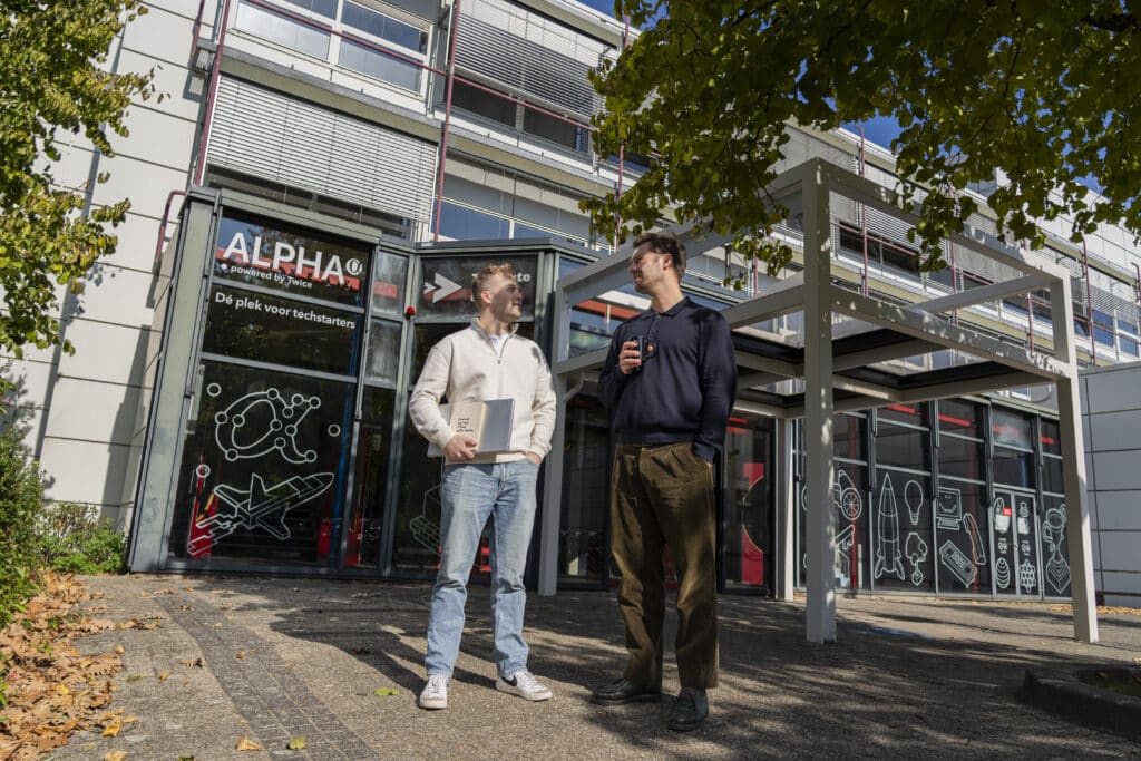 Two people stand outside a modern building with large windows and ALPHA signage. They appear to be talking and holding books or folders. Trees and scattered leaves are visible in the foreground.