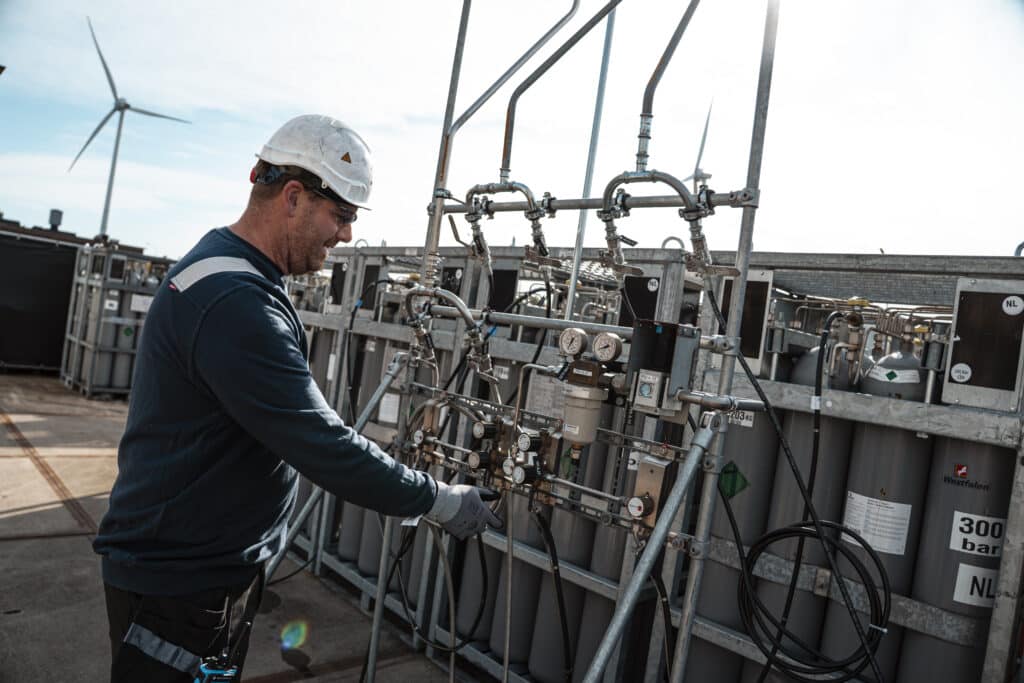 A worker wearing safety gear operates a complex control panel with pipes and gauges outdoors at an industrial site; wind turbines are visible in the background.