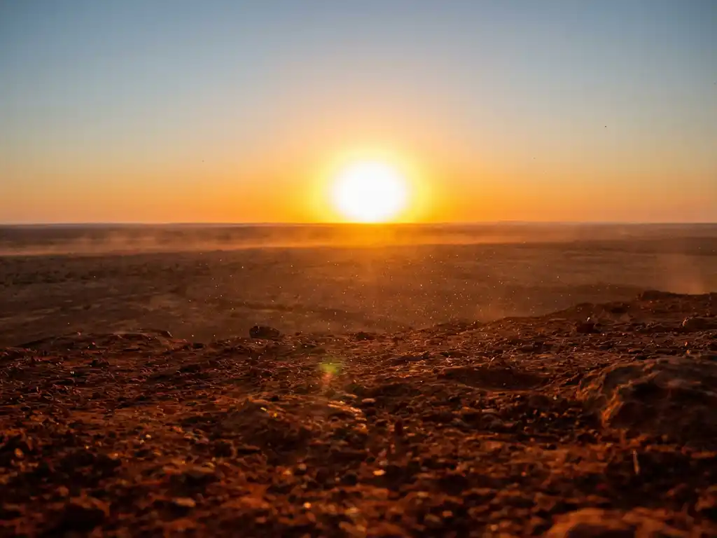 Massive golden sun over iron-red desert landscape, warm amber light illuminating copper dust particles drifting through hazy air.