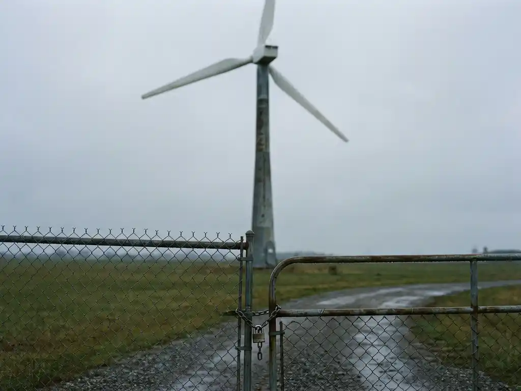 Wind turbine behind a padlocked chain-link fence on a gravel path, surrounded by open fields under an overcast sky.