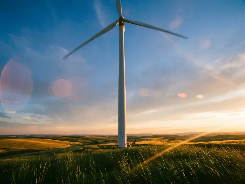 Wind turbine rising from green farmland at golden hour, three white blades mid-rotation against a dramatic amber-lit sky.